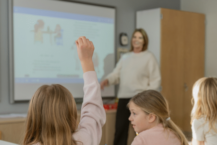 Lärare undervisar en klass framför en interaktiv skrivtavla, en elev räcker upp handen